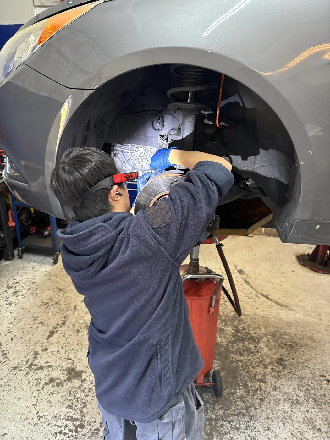 RepairTech Automotive technician in safety glasses and gloves inspecting a front brake rotor with the wheel removed at the Kissimmee shop